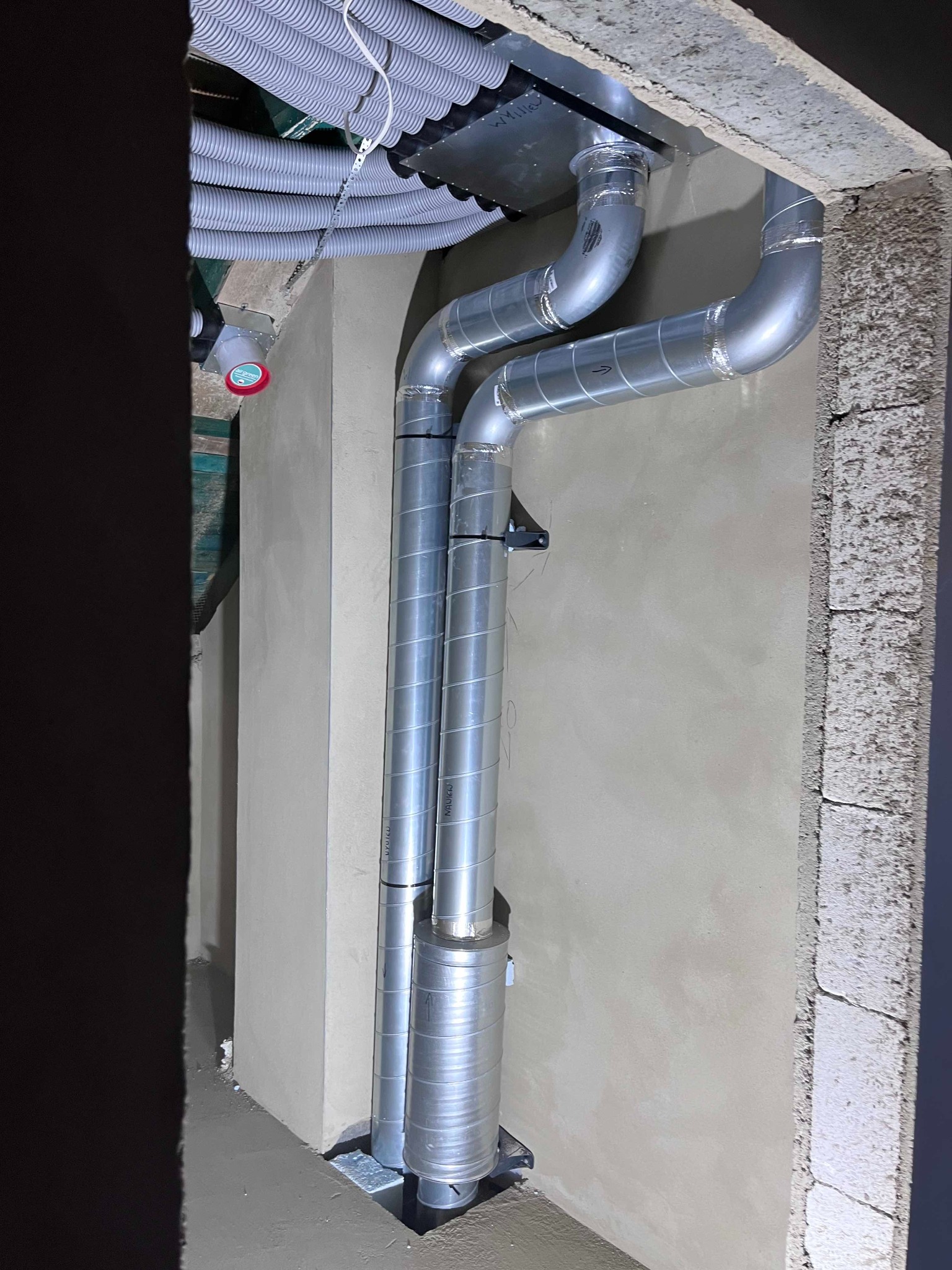 Szare, metalowe rury wentylacyjne, częściowo owinięte taśmą, prowadzone wzdłuż ściany z surowego betonu i świeżego tynku w trakcie montażu systemu wentylacji w pomieszczeniu.