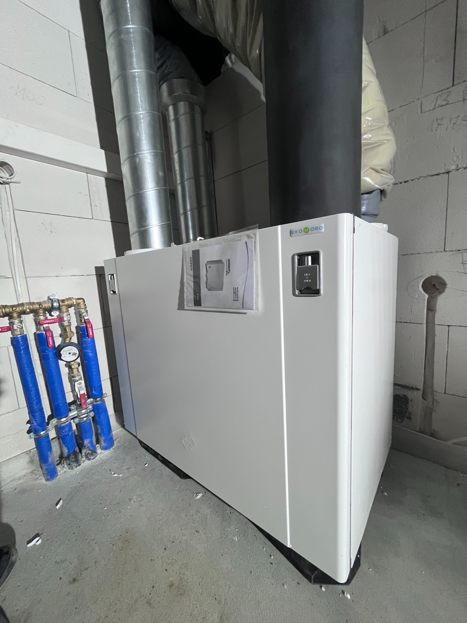 Biała jednostka centralna wentylacyjna Ekonord z podłączonymi rurami wentylacyjnymi i instalacją hydrauliczną, zamontowana przy ścianie z bloczków betonowych.