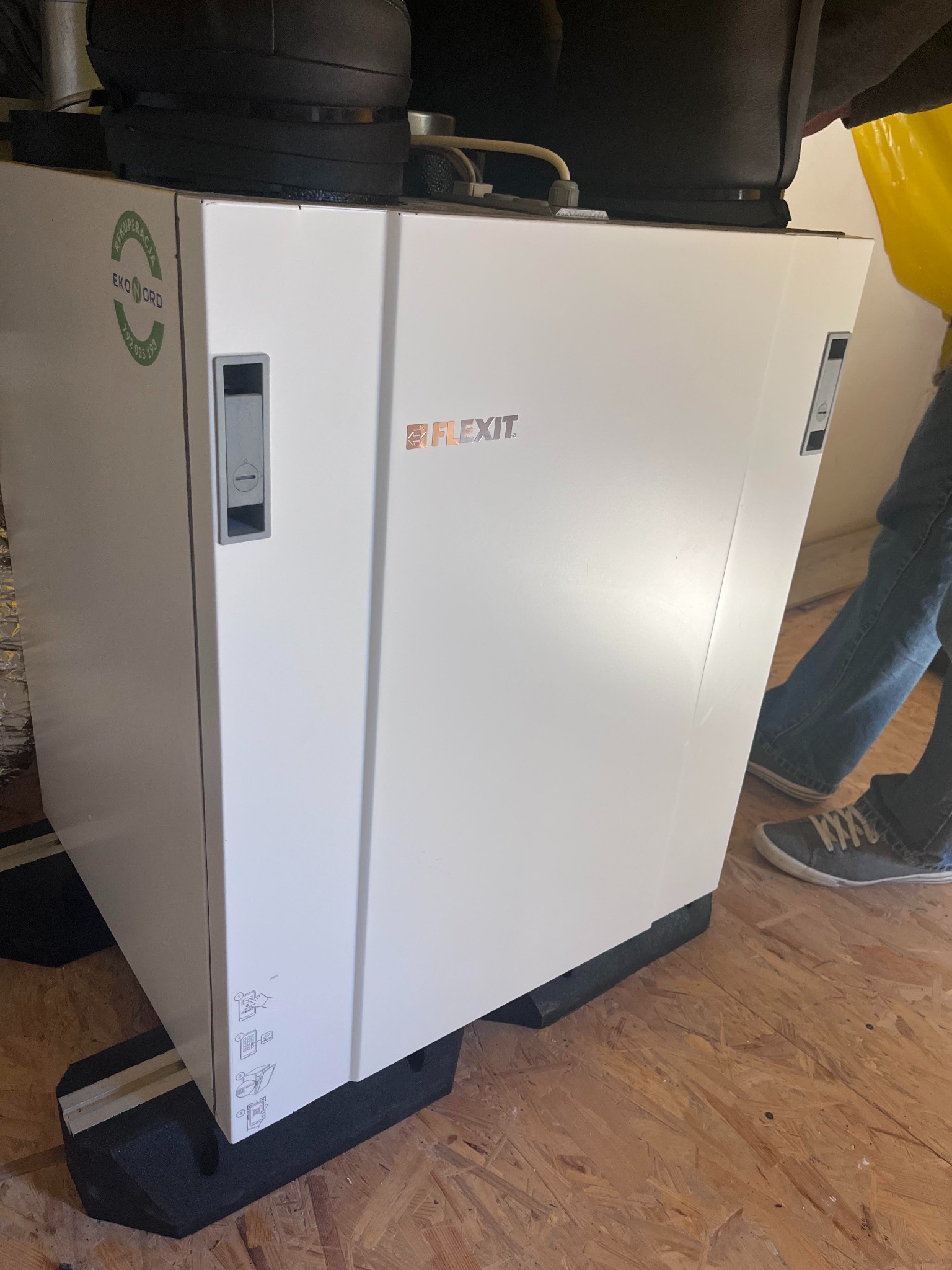 Biała jednostka rekuperacyjna marki Flexit z widocznym logo i naklejką Eko Nord, umieszczona na czarnych podkładkach na podłodze z płyt OSB, z widocznymi elementami instalacji u góry i fragmentem...