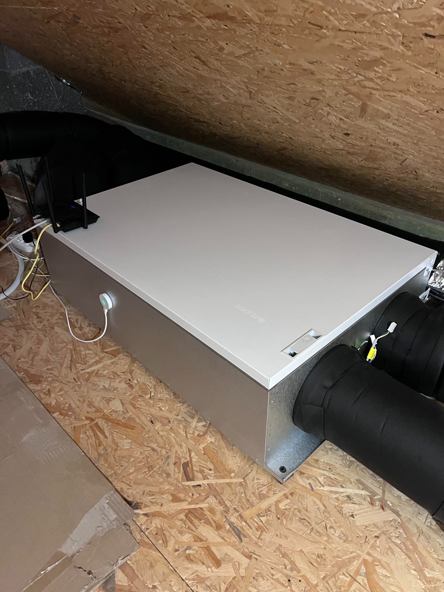 Centrala wentylacyjna z podłączonymi przewodami, umieszczona na poddaszu z boazerią, widoczny router WiFi obok urządzenia.