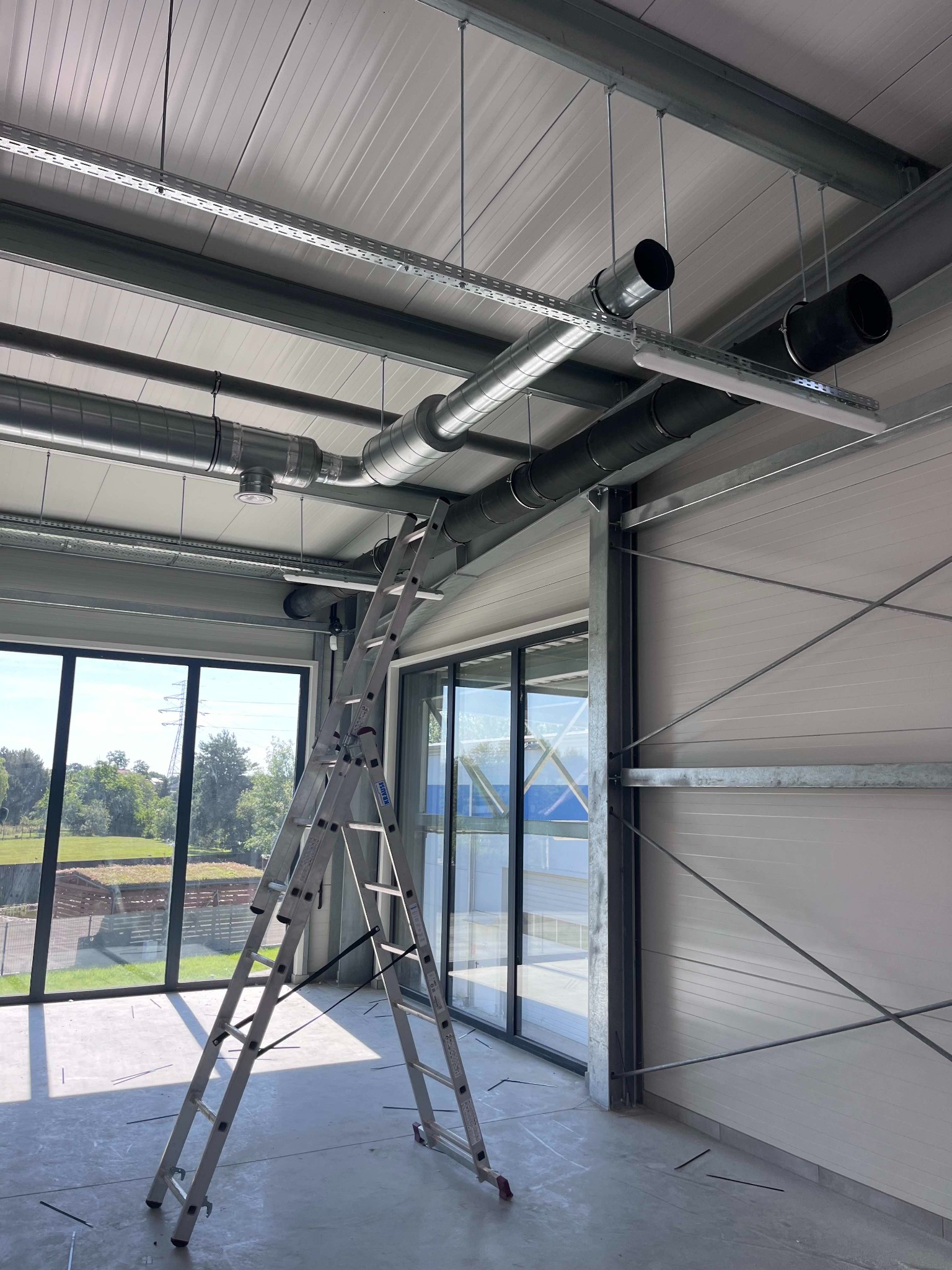 Wnętrze hali przemysłowej z widocznym systemem rur wentylacyjnych pod sufitem oraz rozstawioną drabiną Krause na tle dużych okien.