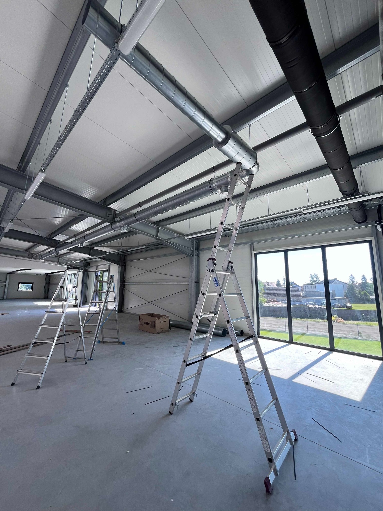 Wnętrze hali przemysłowej w trakcie montażu instalacji wentylacyjnej; widoczne drabiny, kanały wentylacyjne z blachy ocynkowanej i czarne, izolowane rury wentylacyjne pod sufitem, okna z widokiem...