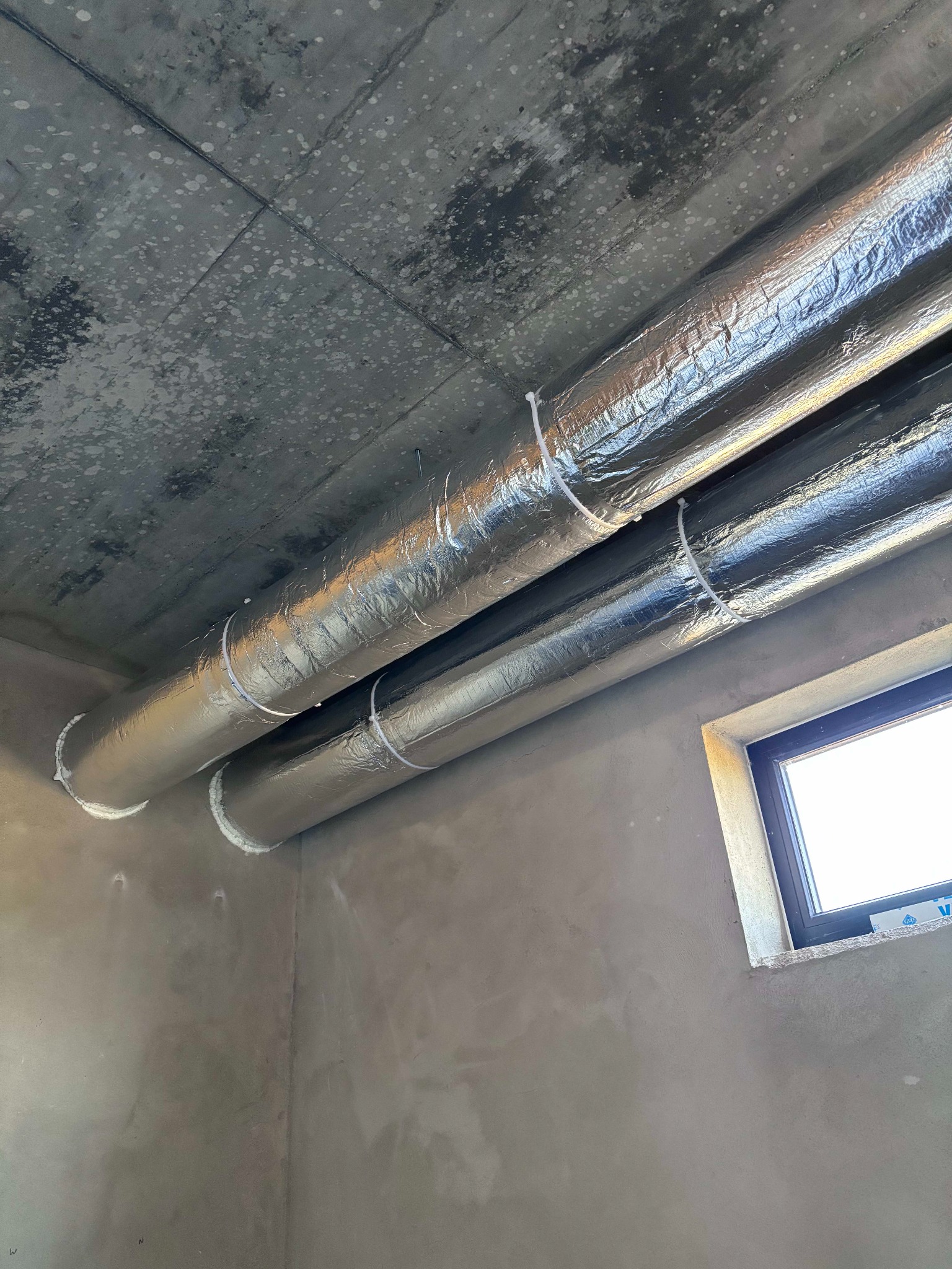 Dwa izolowane przewody wentylacyjne zamontowane pod surowym, betonowym sufitem, zabezpieczone białymi opaskami zaciskowymi i uszczelnione pianką montażową przy wlotach, obok widoczne okno.