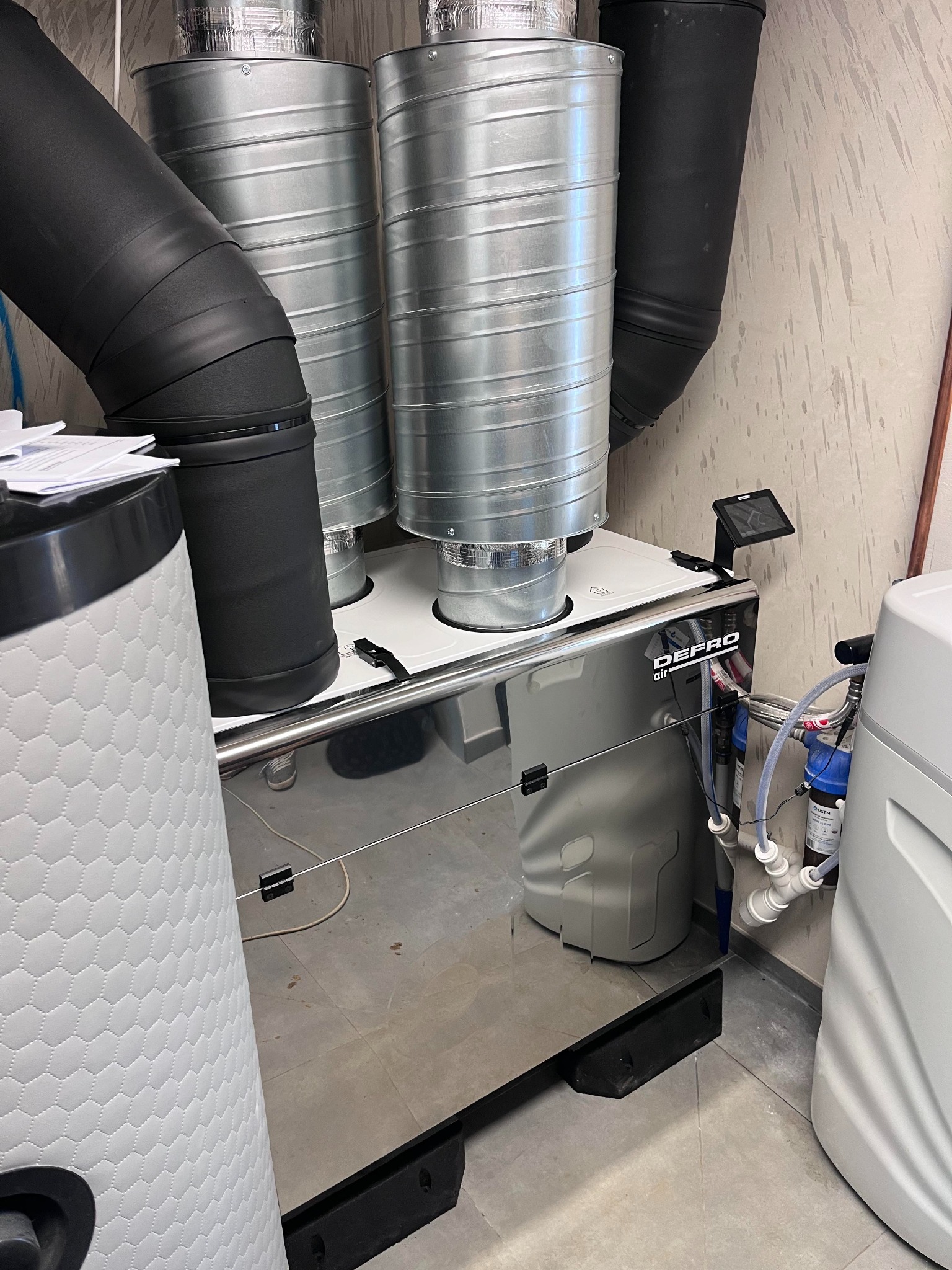 Srebrny rekuperator marki DEFRO z czarnymi i srebrnymi przewodami wentylacyjnymi, widoczny tablet sterujący oraz elementy instalacji wodnej w tle.