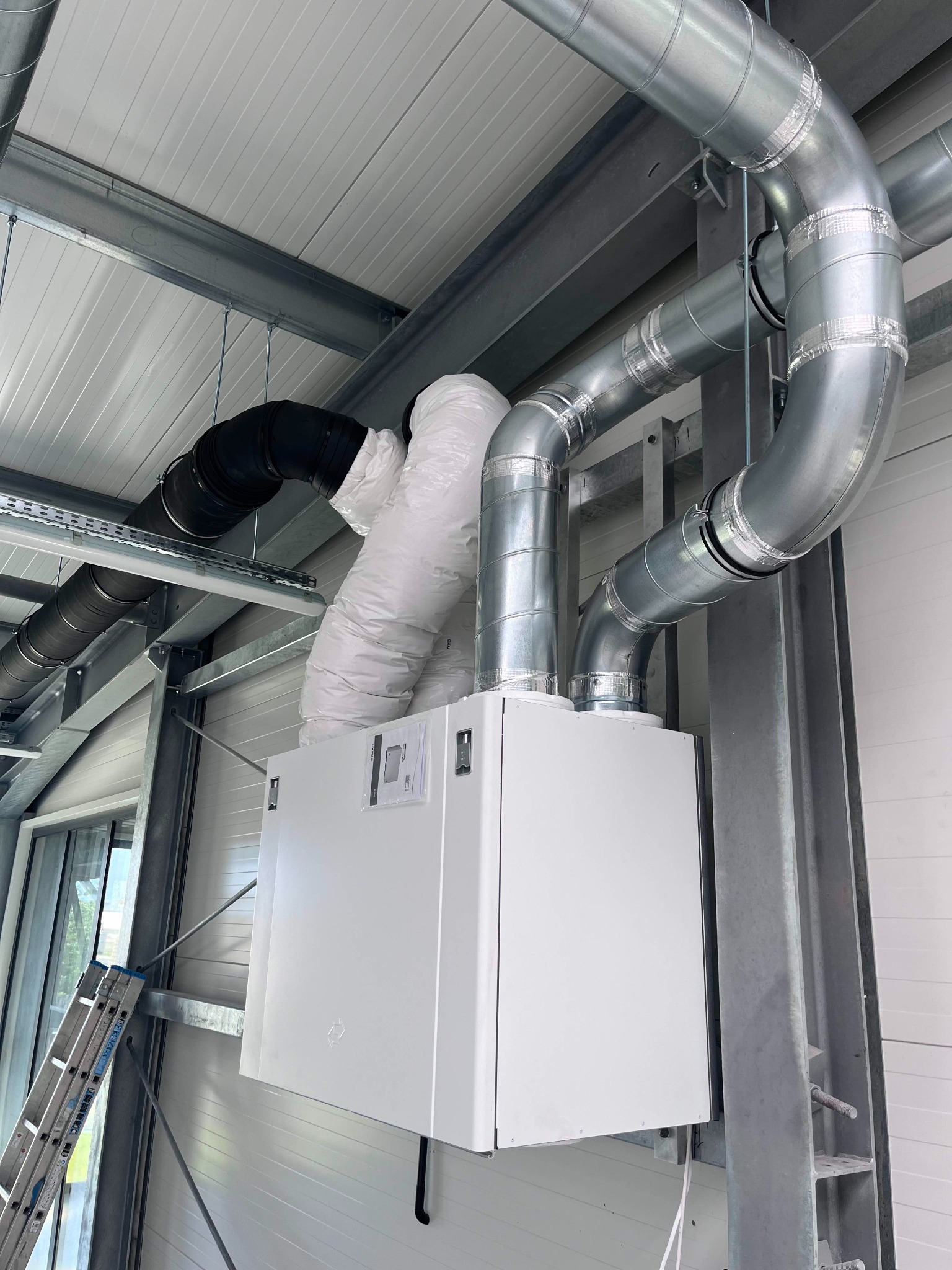 Biała centrala wentylacyjna z podłączonymi przewodami wentylacyjnymi w budynku o metalowej konstrukcji, widoczna drabina oparta o ścianę.