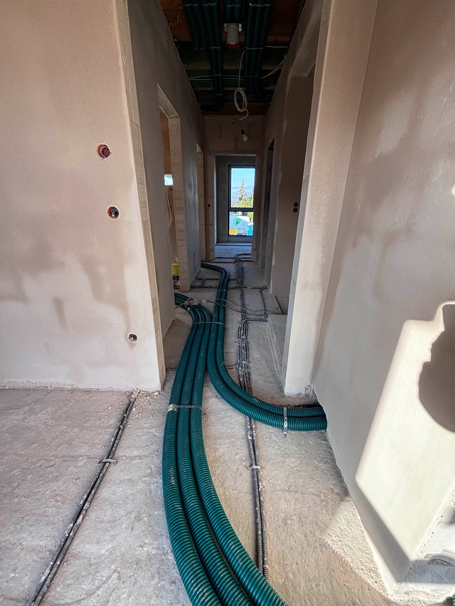 Wnętrze w stanie surowym z widocznymi, zielonymi rurami wentylacyjnymi ułożonymi na podłodze, prowadzącymi wzdłuż korytarza. Na ścianach widoczne puszki elektryczne. Rury przymocowane do podłoża...