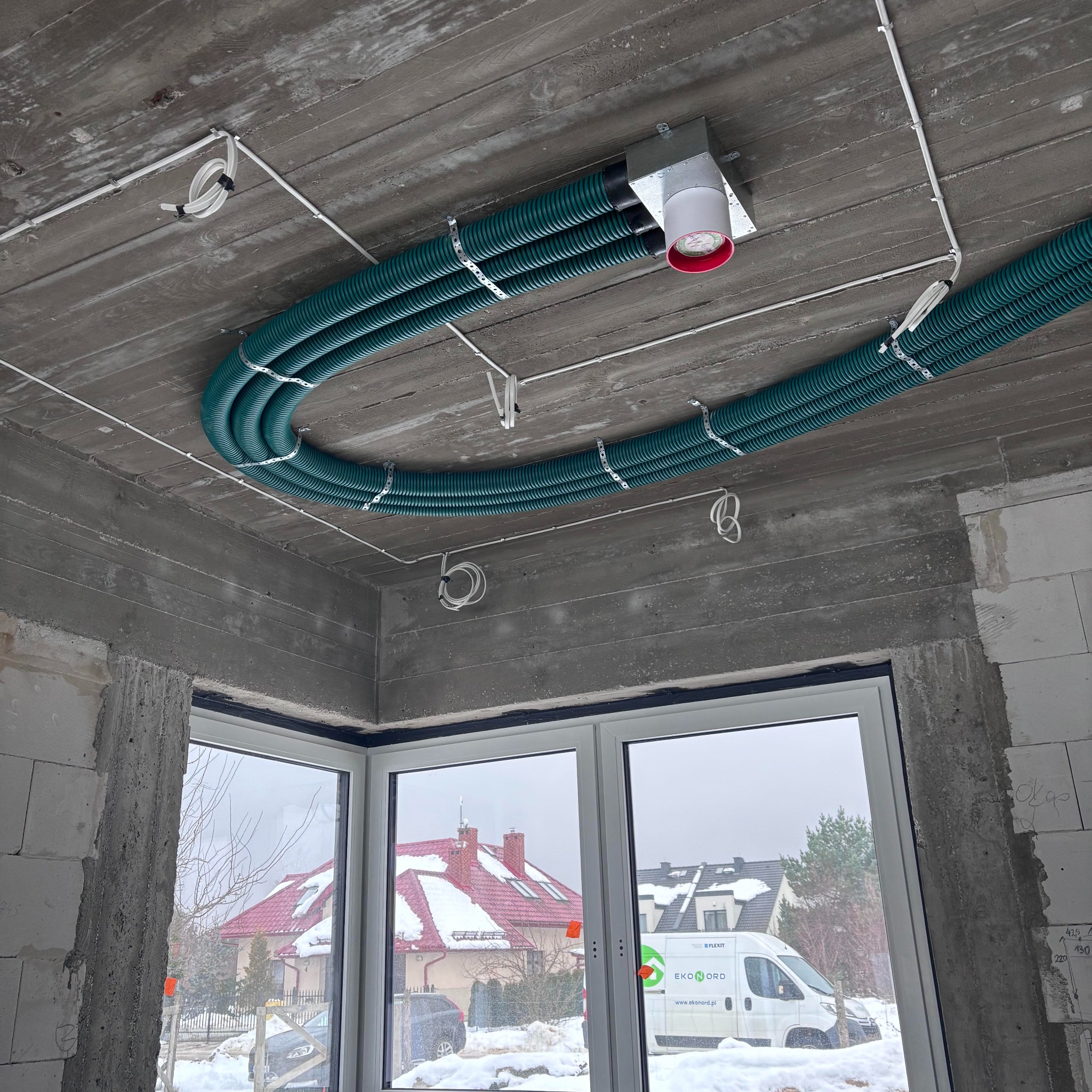 Niedokończona instalacja wentylacyjna z trzema zielonymi, karbowanymi rurami przymocowanymi do surowego betonowego sufitu za pomocą metalowych obejm, widoczny element centralny z czerwoną obwódką...