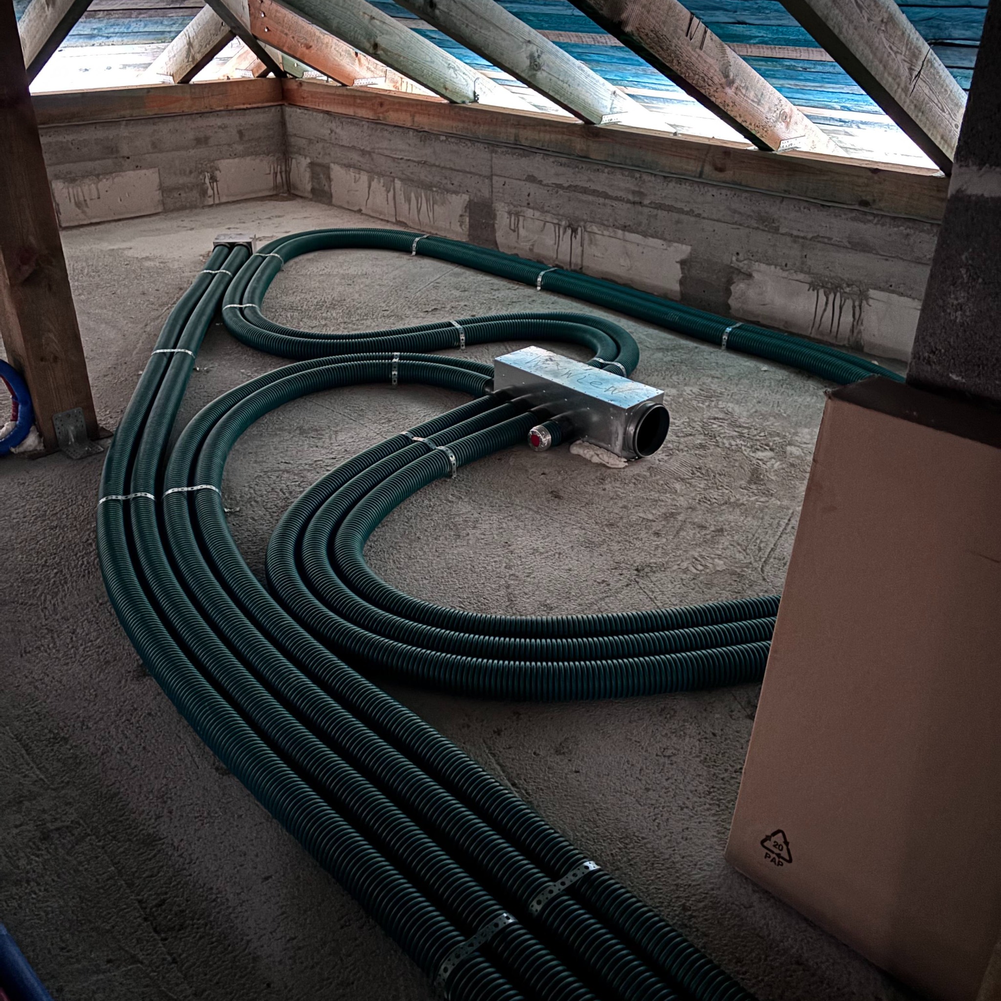 Wnętrze poddasza z widocznym systemem rur wentylacyjnych, połączonych metalową skrzynką rozdzielczą, z widoczną konstrukcją dachu i fragmentem kartonu.