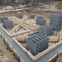 Wykop pod fundamenty z drewnianym szalunkiem wypełnionym świeżym betonem, na którym ustawione są bloczki betonowe. Widoczne ślady opon i błoto. W tle drzewa bez liści.