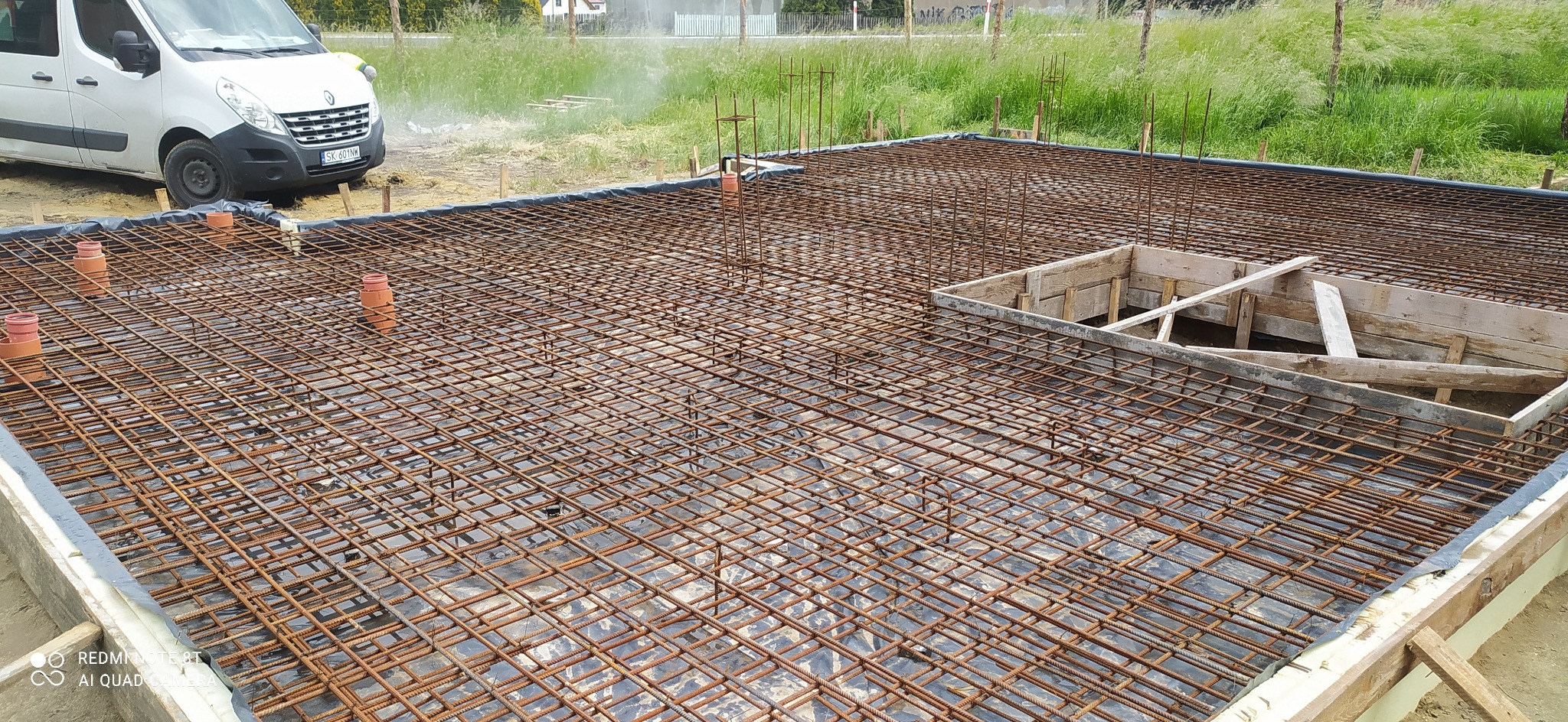Zbrojenie fundamentu z siatki stalowej na czarnej folii, z fragmentem szalunku z desek, widok z góry w trakcie prac budowlanych, z zaparkowanym białym samochodem dostawczym w tle.