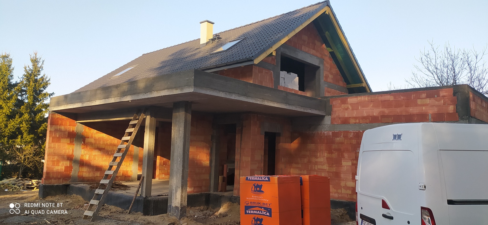 Budowa domu w stanie surowym otwartym, widoczne ściany z czerwonej cegły, betonowe słupy i strop, drewniana drabina oparta o budynek, dach pokryty dachówką, palety z bloczkami Termalica obok...