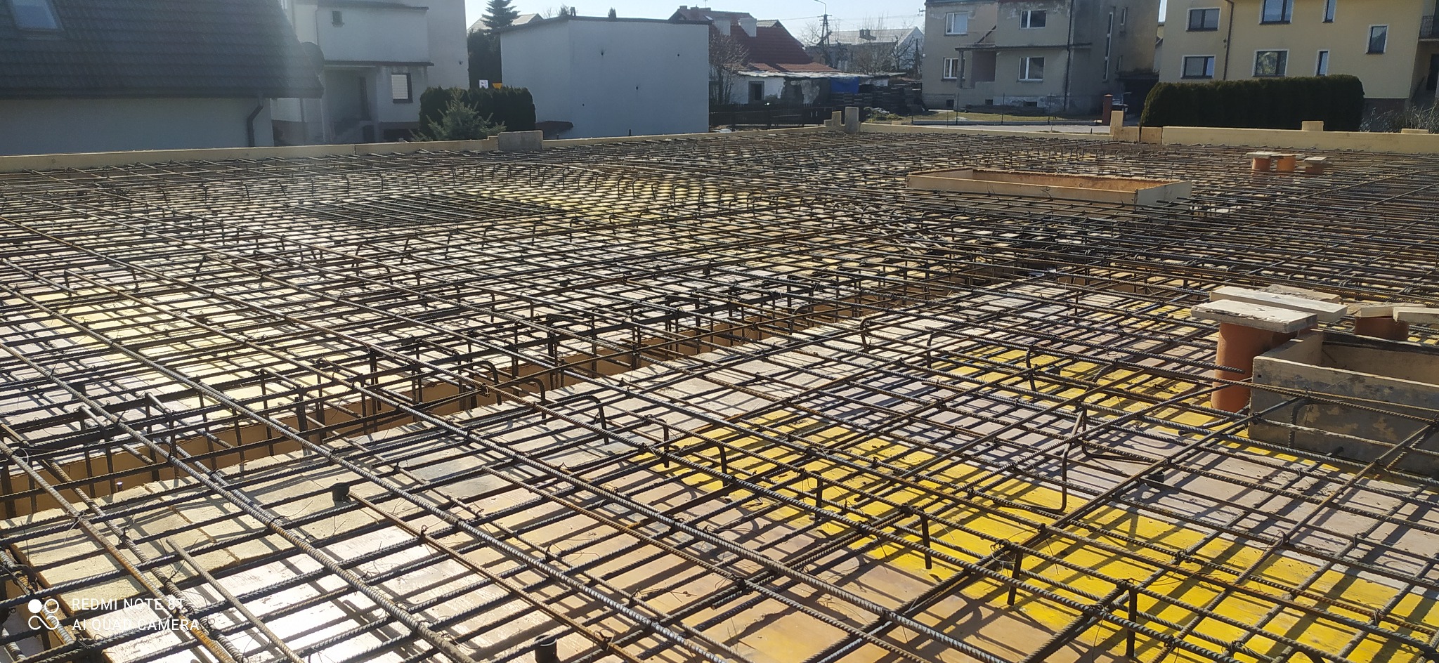 Siatka zbrojeniowa ułożona na żółtym szalunku, przygotowana do zalania betonem, widok z góry na tle zabudowań.