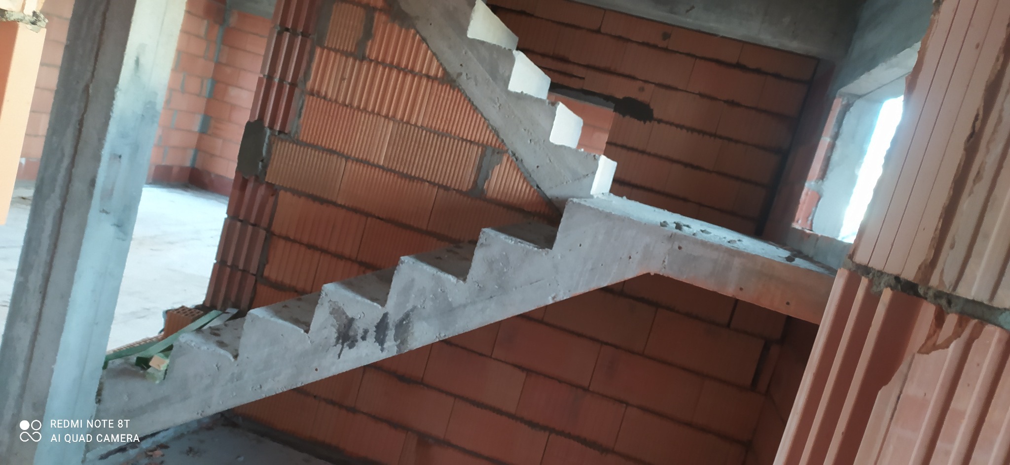 Surowe, betonowe schody wewnętrzne w trakcie budowy domu z czerwonej cegły, widoczne niedoskonałości i chropowata faktura betonu, surowy stan wykończenia wnętrza.