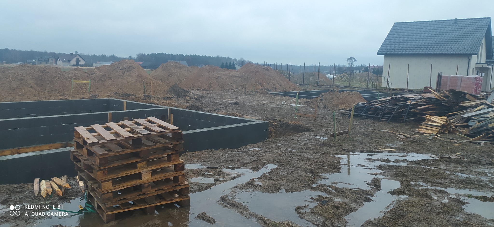 Wykop pod fundamenty domu jednorodzinnego, z widocznymi zalanymi betonem ścianami szalunkowymi, stertą desek i paletą na pierwszym planie, w tle hałdy ziemi i budynki w oddali, pochmurny dzień.