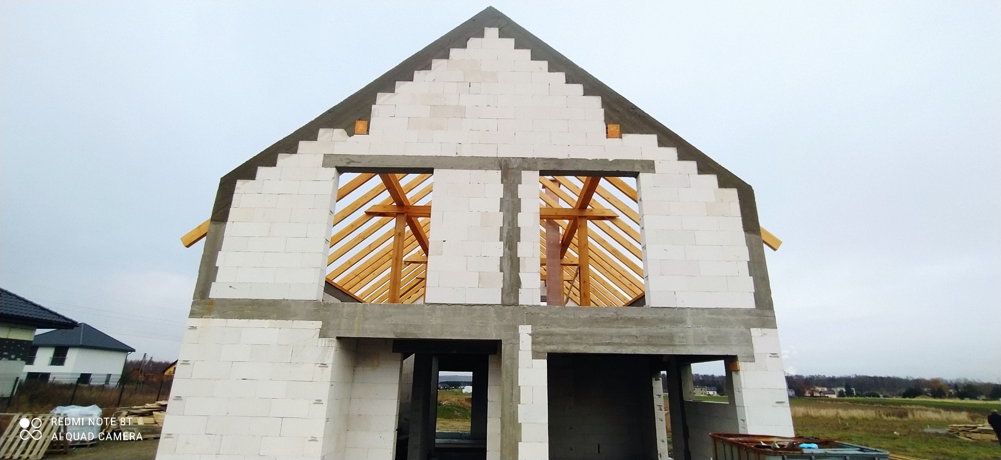Surowy stan budynku z białych bloczków, widoczne drewniane elementy konstrukcji dachu przez otwory okienne, szare nadproża i filary, w tle pochmurne niebo i zabudowa.