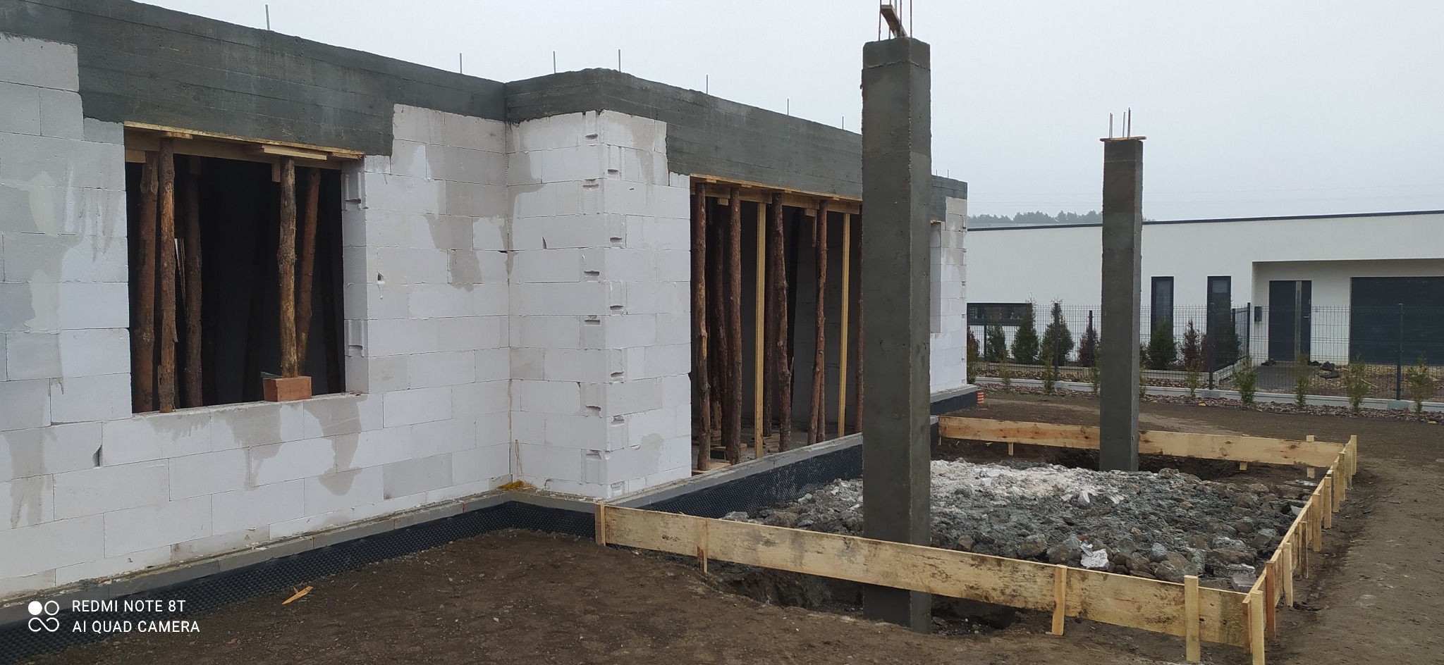 Wznoszenie ścian z betonu komórkowego na fundamencie, widoczne drewniane stemple podtrzymujące otwory okienne, betonowe filary w szalunku, teren budowy przygotowany pod wylewkę.