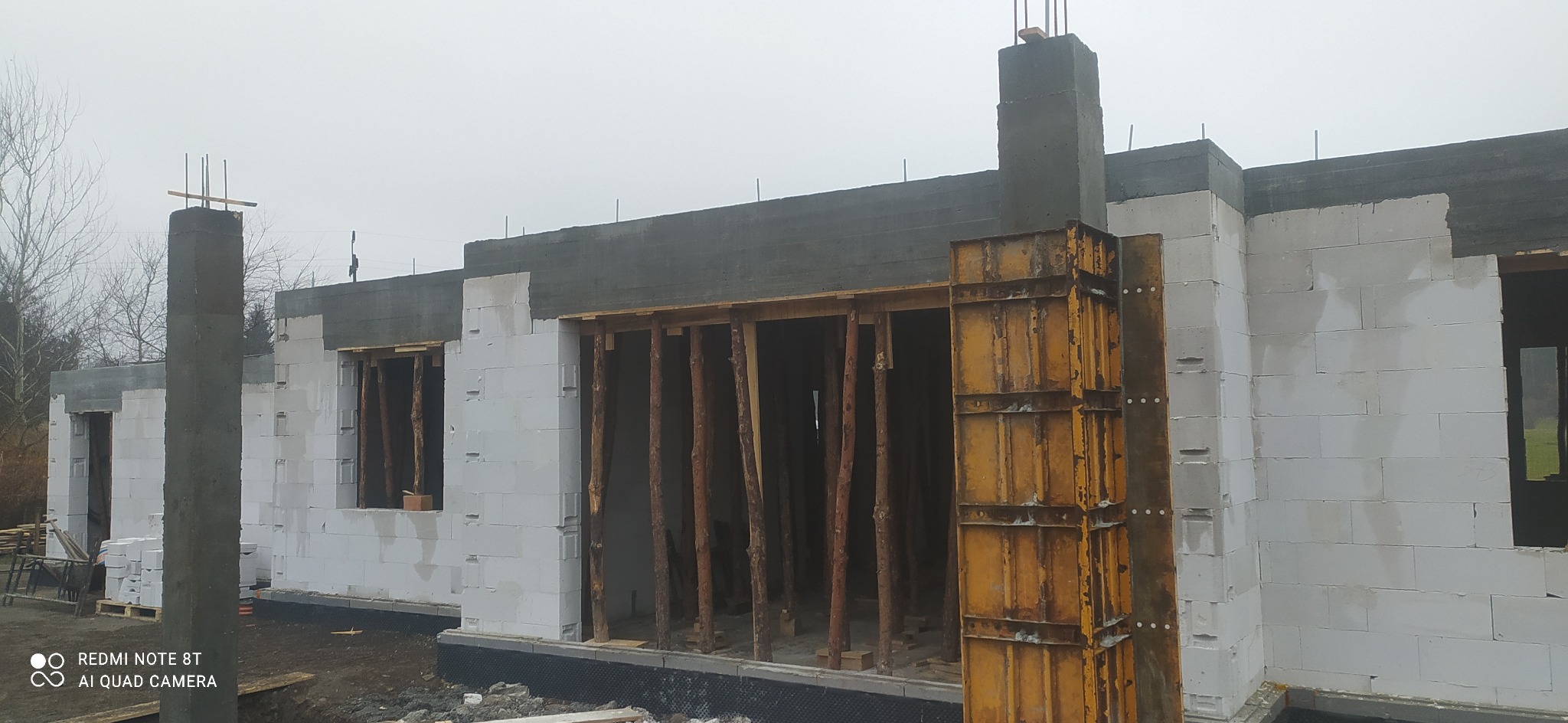 Wznoszenie ścian domu z bloczków, widoczne drewniane stemple podtrzymujące nadproże oraz żółte szalunki narożne, wystające pręty zbrojeniowe.