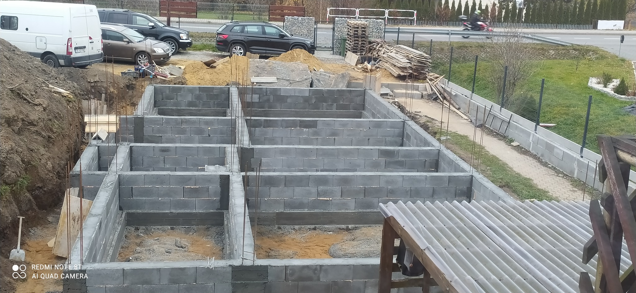 Wykop pod fundamenty z częściowo wzniesionymi ścianami z bloczków betonowych, wystającymi prętami zbrojeniowymi, widok z góry na plac budowy z zaparkowanymi samochodami i stertą drewna w tle.