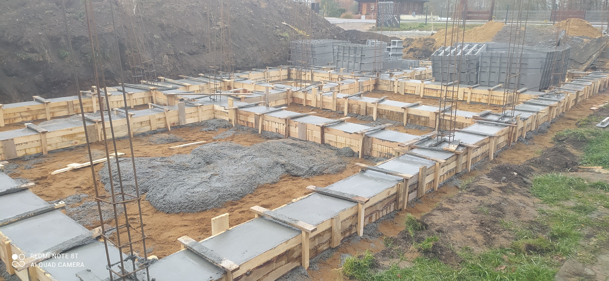 Wylane fundamenty z drewnianym szalunkiem, widoczne zbrojenia i świeży beton, ziemia wokół.