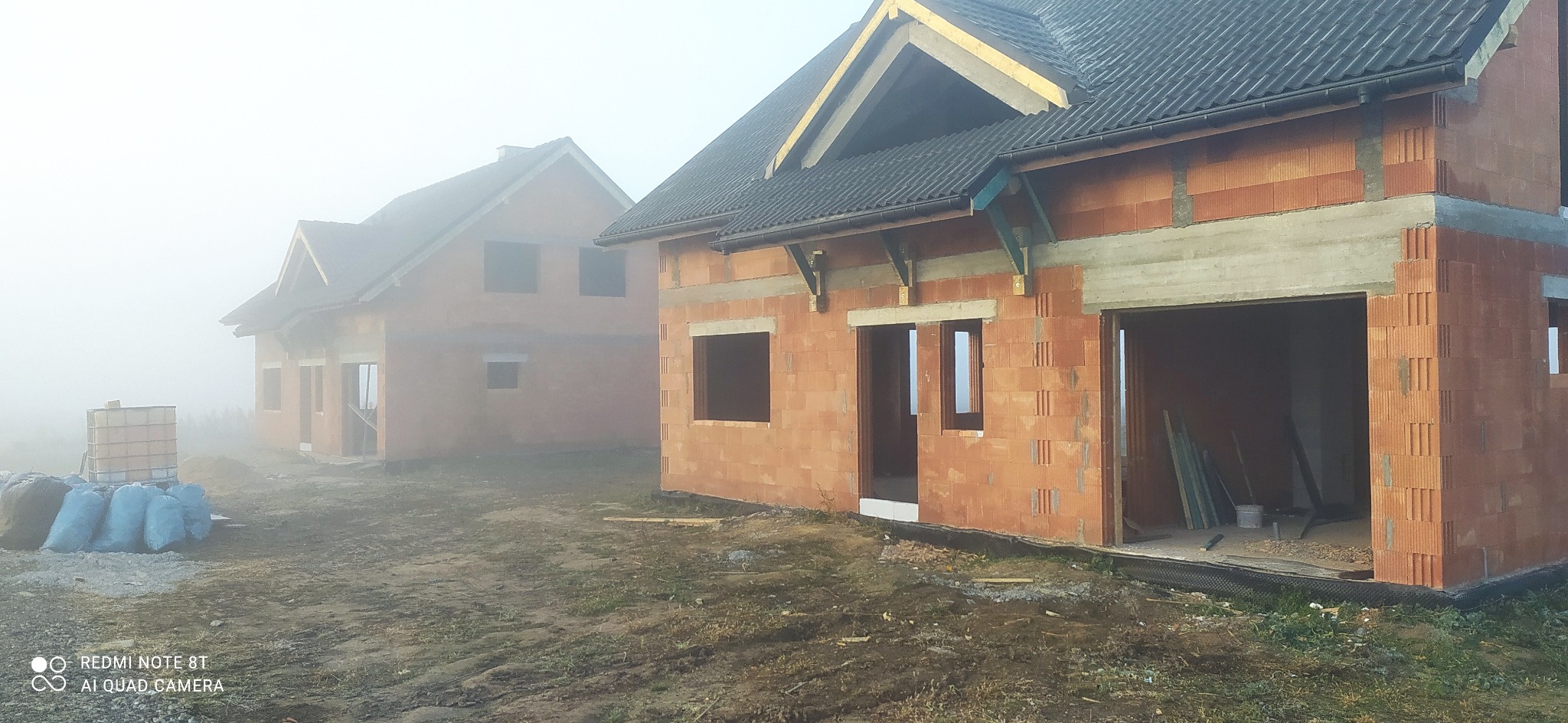 Dwa budynki w stanie surowym otwartym z czerwonej cegły ceramicznej, z widocznym zadaszeniem i przygotowanymi otworami okiennymi, spowite poranną mgłą.