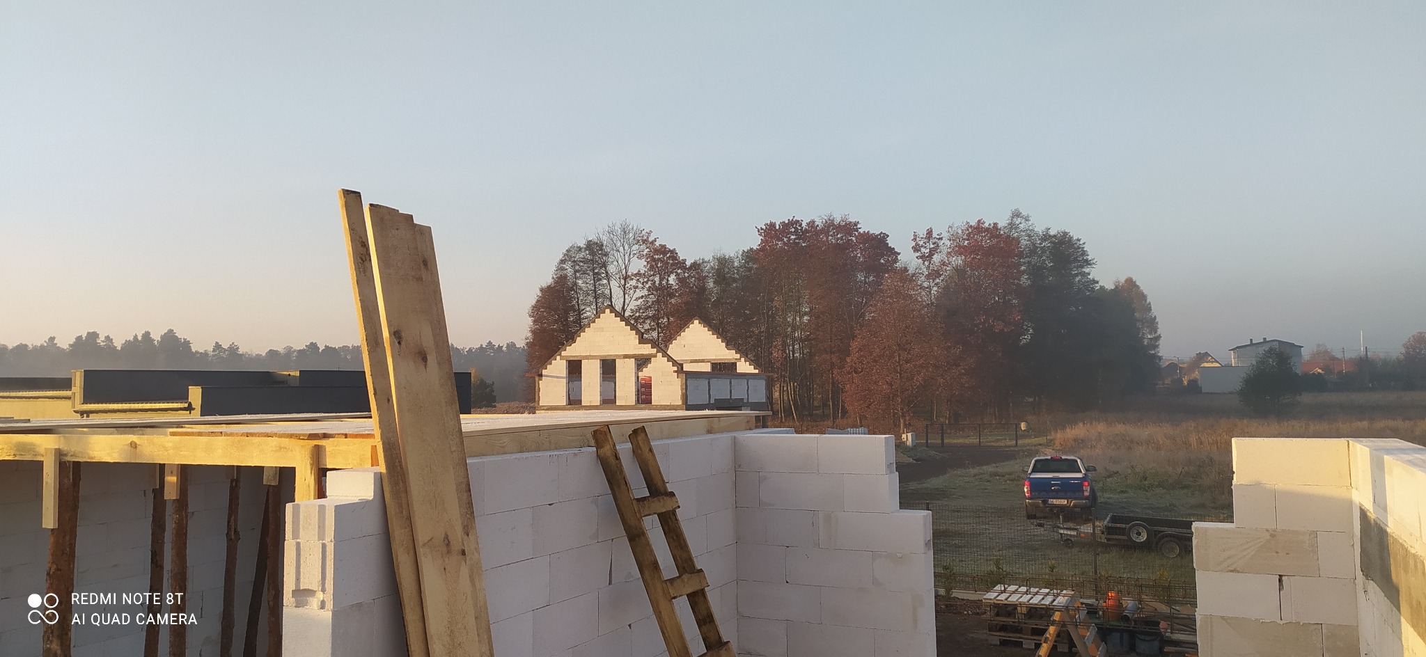 Wczesnoporanny widok na plac budowy domu z bloczków, z widocznymi drewnianymi elementami szalunku i drabiną opartą o ścianę, w tle dwa budynki w stanie surowym otoczone drzewami i lekka mgła.