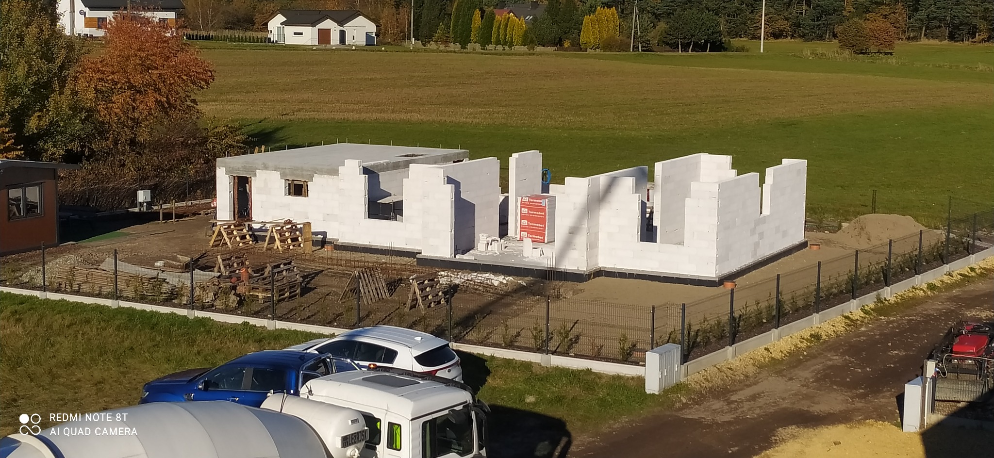 Wznoszenie ścian parteru z bloczków betonu komórkowego na fundamencie, widoczne zbrojenie, palety z bloczkami, w tle zabudowa jednorodzinna i zieleń.