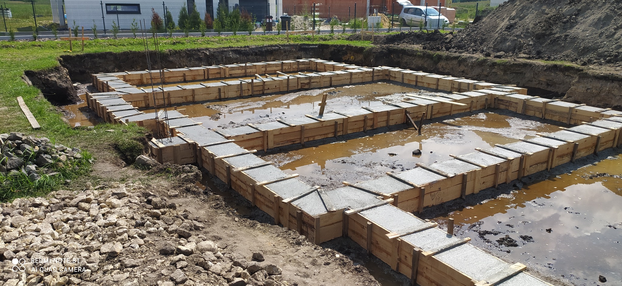 Wykop pod fundamenty z drewnianym szalunkiem wypełnionym świeżym betonem, częściowo zalany wodą, widok z góry na plac budowy.