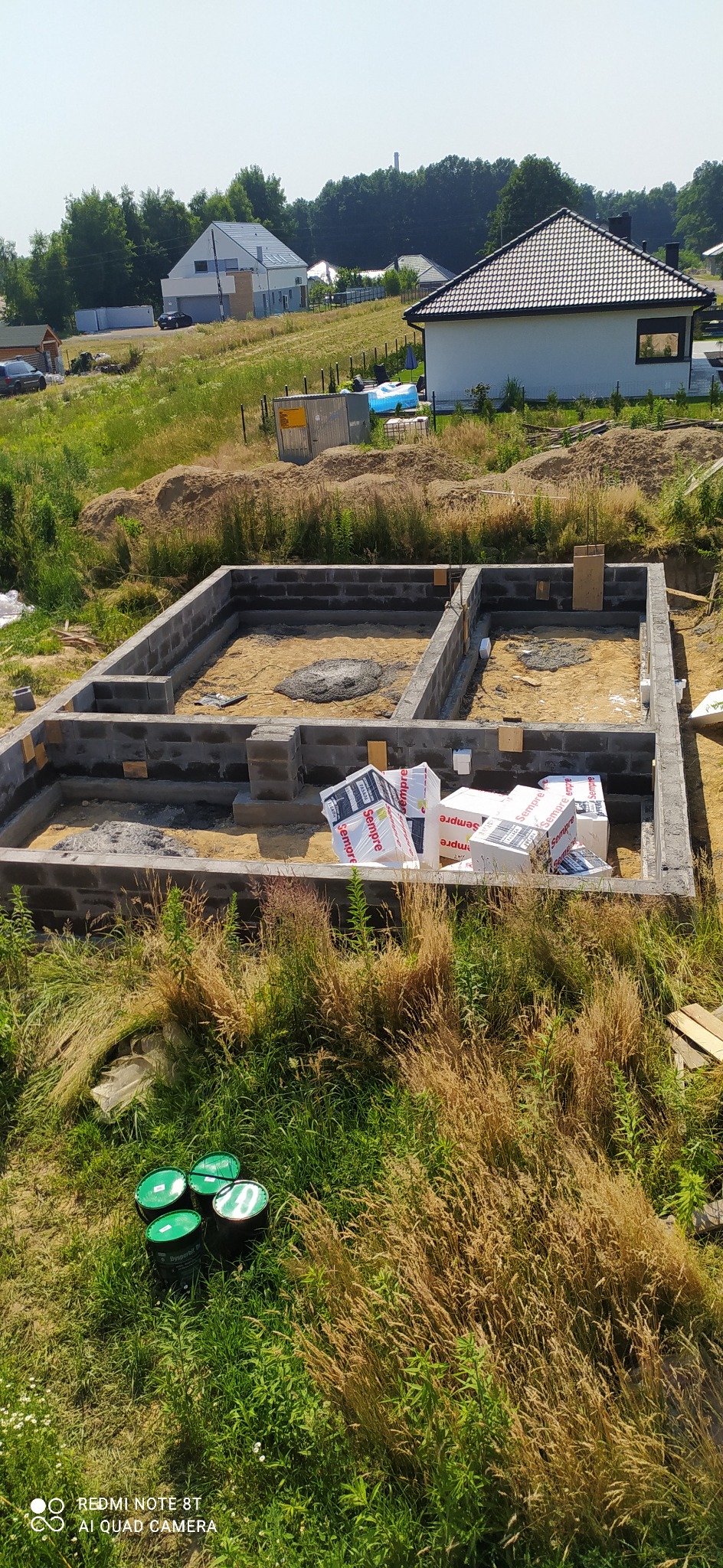 Wykop pod fundamenty domu jednorodzinnego z widocznymi bloczkami betonowymi, częściowo wypełniony piaskiem, z izolacją termiczną w opakowaniach i zielonymi pojemnikami na pierwszym planie.