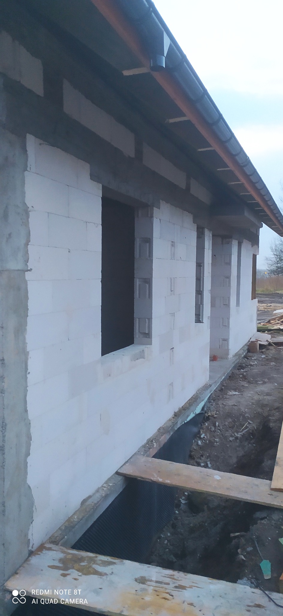 Ściana domu murowanego w trakcie budowy, widoczne otwory okienne, ściana z białych bloczków, zabezpieczona fundamentowa izolacja pionowa, częściowo odsłonięty fundament.