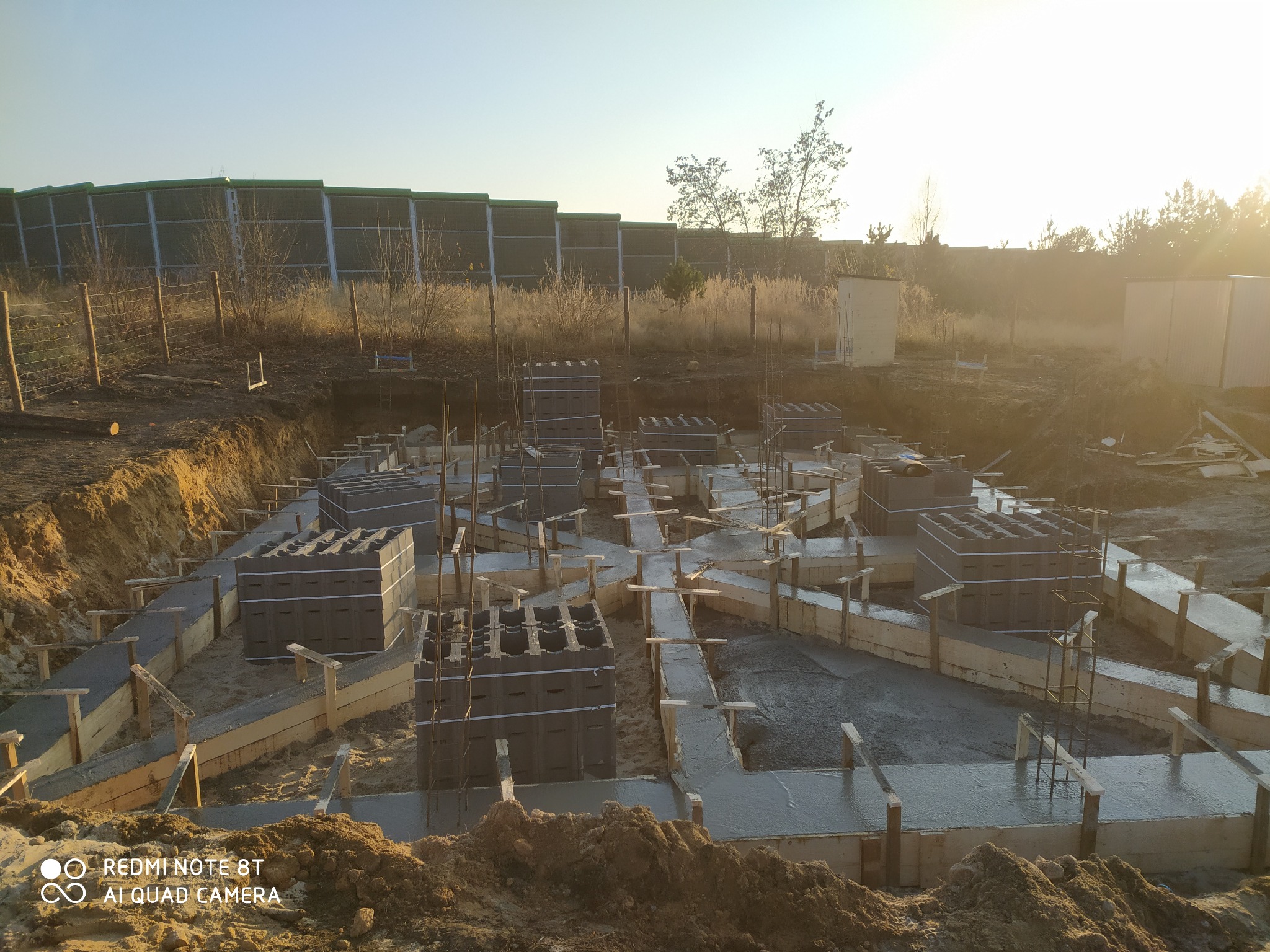 Wykop pod fundamenty z częściowo wylanym betonem w szalunkach, widoczne bloczki betonowe i wystające pręty zbrojeniowe, w tle ziemia, trawa i płot akustyczny.