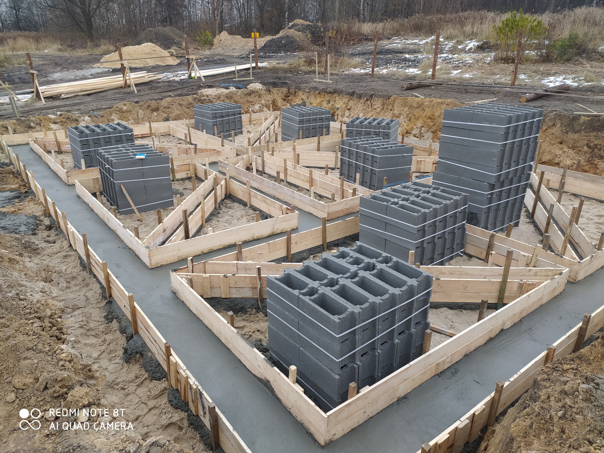 Wykop pod fundamenty z drewnianym szalunkiem wypełnionym świeżym betonem, na którym ustawione są bloczki betonowe. Widoczne ślady opon i błoto. W tle drzewa bez liści.