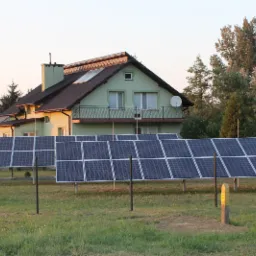 Panele słoneczne na trawiastym terenie przed domem z balkonem, widoczne podpory paneli i częściowo zmontowane konstrukcje w tle.