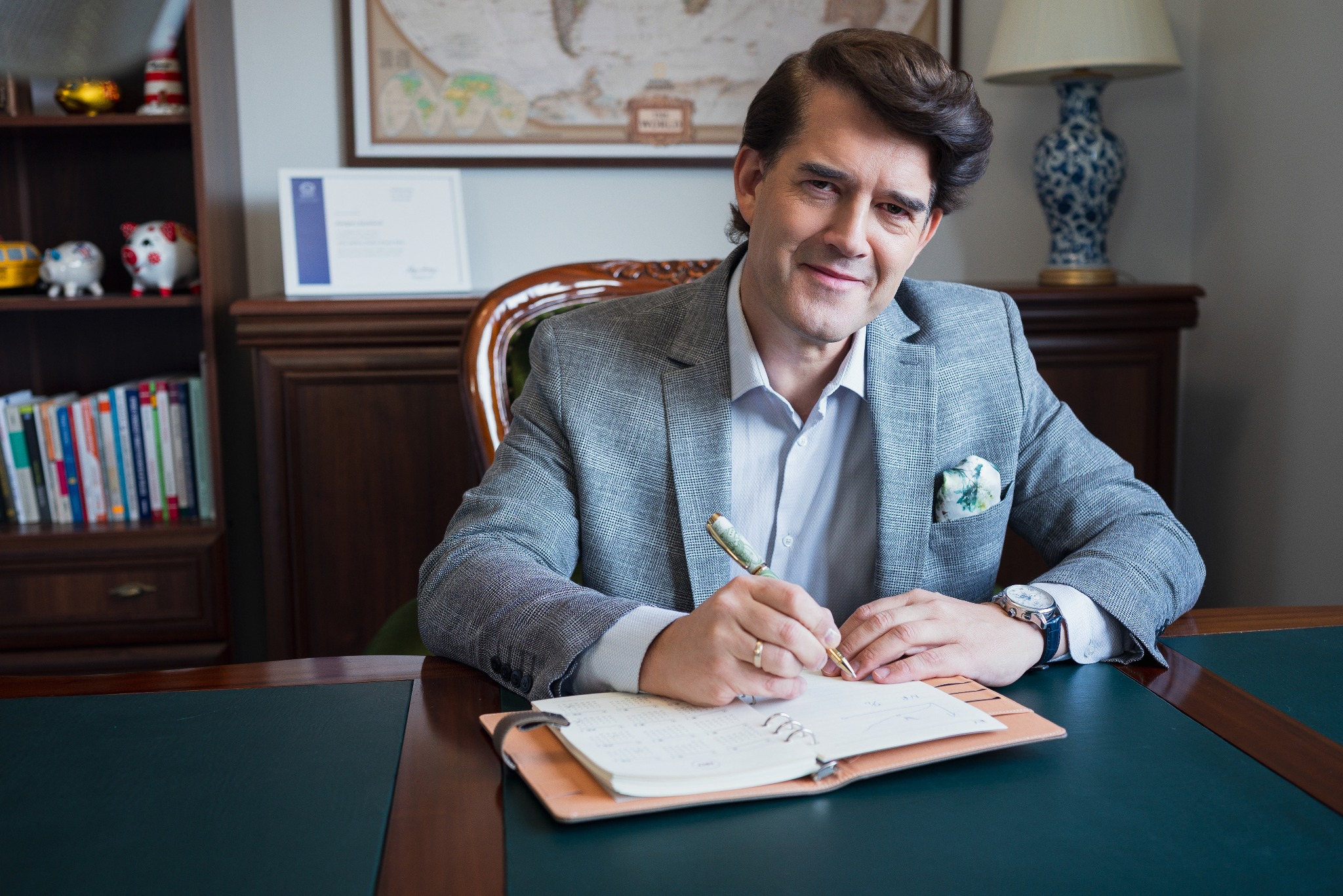 Mężczyzna w tweedowej marynarce notuje w kalendarzu przy biurku. W tle regał z książkami i mapa świata. Elegancki długopis i zegarek dodają profesjonalizmu.