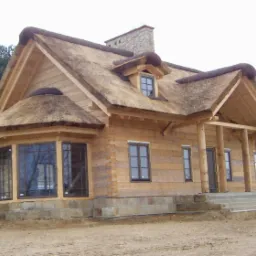 Drewniany dom szkieletowy z unikalnym strzechowym dachem i kamiennym kominem. Widoczna weranda z drewnianymi kolumnami i okna z ciemnymi ramami.