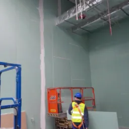 Praca przy montażu płyt karton-gips na ścianie w dużym pomieszczeniu. Widoczny pracownik na podnośniku, szpachlowane łączenia. Instalacje pod sufitem.