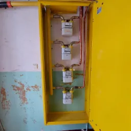 Cztery liczniki gazu w żółtej szafce, z miedzianymi rurami, na tle ściany z odpryskującą farbą i kafelkami na podłodze.