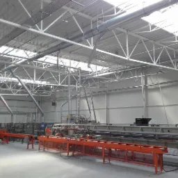 Wnętrze hali produkcyjnej z widocznymi instalacjami wentylacyjnymi pod sufitem i linią produkcyjną. Metalowa konstrukcja dachu z oknami. Szare ściany.