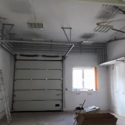 Wnętrze hali przemysłowej z widoczną bramą segmentową, instalacjami pod sufitem, drabina aluminiowa oparta o ścianę. Widoczne rury instalacyjne podwieszone pod sufitem oraz oświetlenie rastrowe.