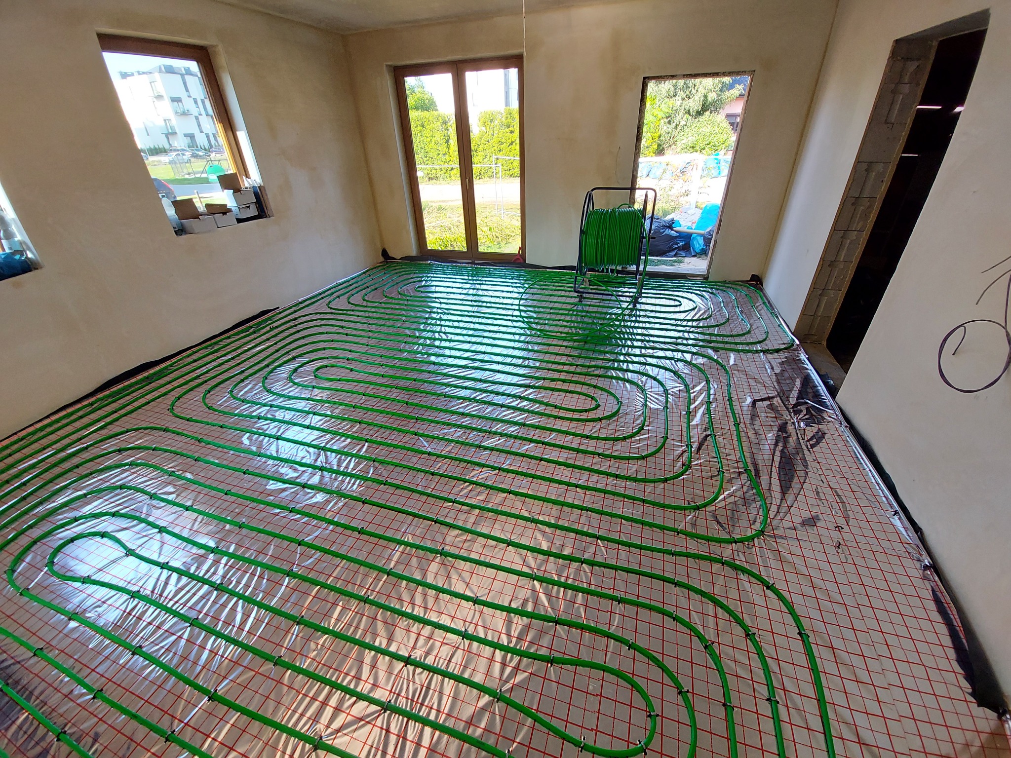 Zielone rury ogrzewania podłogowego rozłożone na izolacji z siatką w surowym wnętrzu, widoczny rozwijak do rur i okna z widokiem na zieleń.