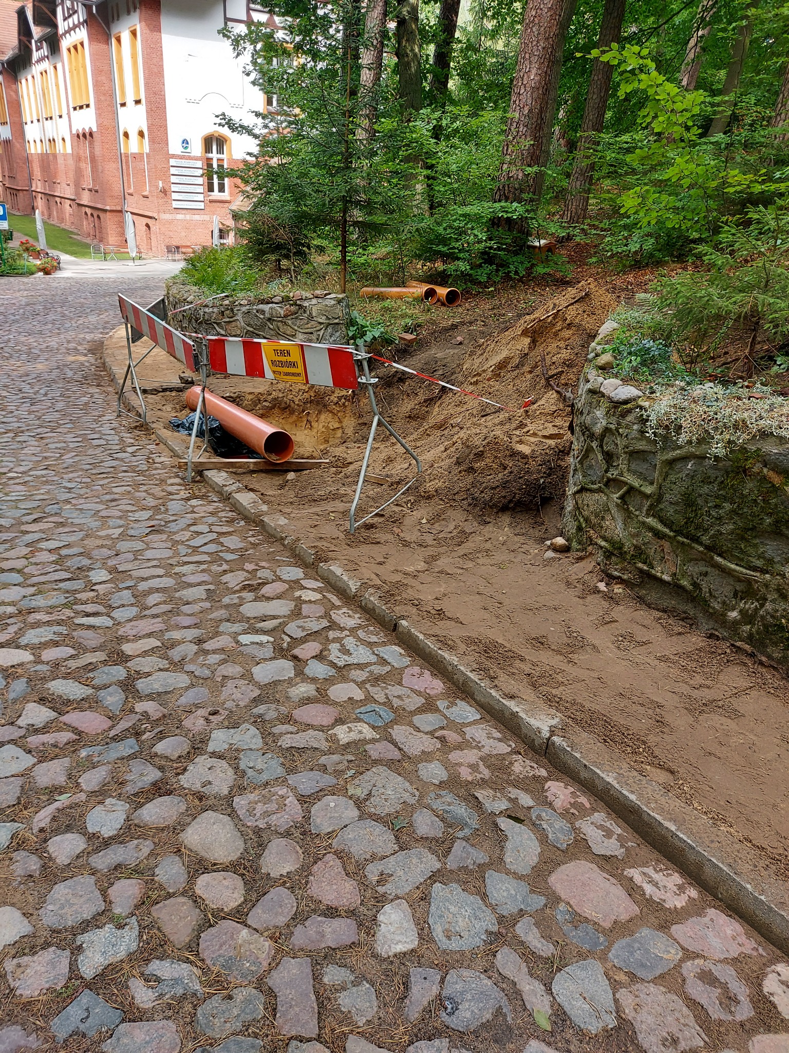 Wykonanie wykopu pod pomarańczowe rury kanalizacyjne w pobliżu kamienicy i brukowanej drogi, teren zabezpieczony barierkami z ostrzeżeniem.