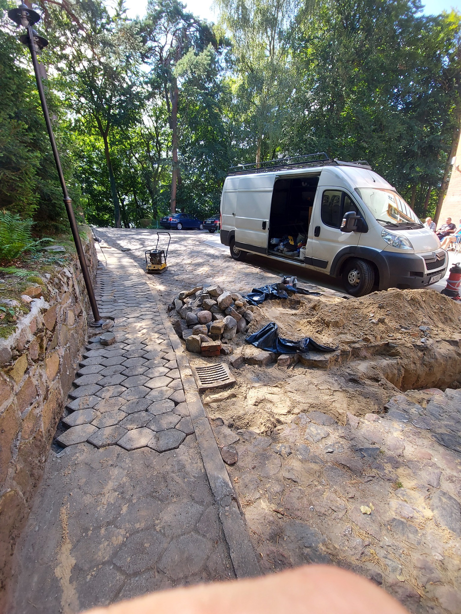Ułożona część chodnika z kostki brukowej, wykopana ziemia, przygotowane kamienie i samochód dostawczy z otwartymi drzwiami w tle sugerują prace ziemne i brukarskie.