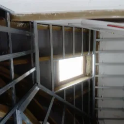 Szkielet metalowy pod zabudowę poddasza, widoczne drewniane krokwie, okno dachowe i częściowo wypełniona wełną mineralną przestrzeń między profilami.