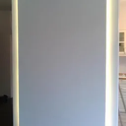 Biała ściana z wbudowanym oświetleniem LED w pionowych szczelinach, widoczny fragment ciemnej podłogi i fragment kuchni w tle.