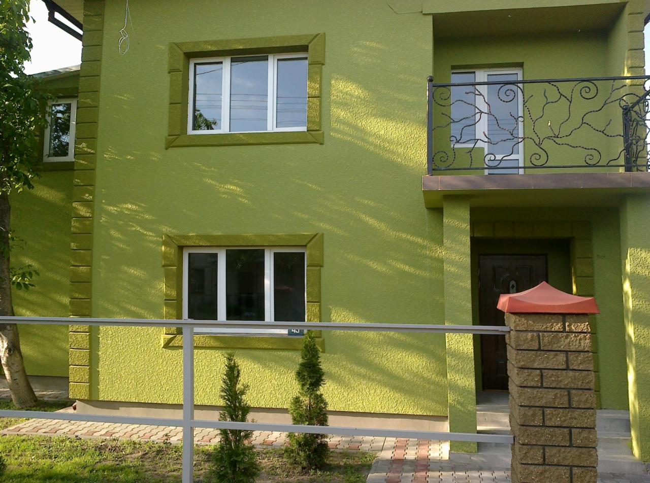 Elewacja domu w odcieniach zieleni z oknami PCV, balkonem z ozdobną, metalową balustradą i ceglanym słupkiem ogrodzenia z czerwonym daszkiem.