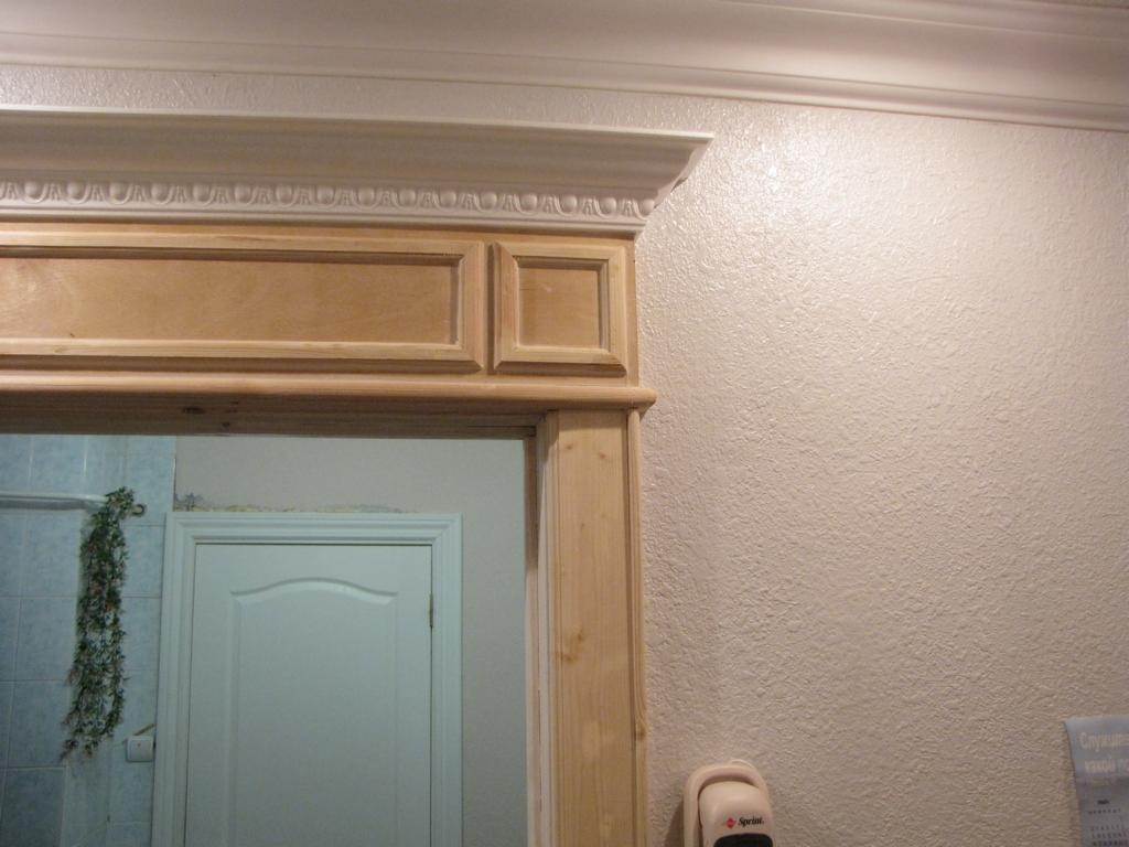 Fragment łazienki z drewnianą obudową drzwi, widoczna biała ściana z teksturą i listwa sufitowa. Po lewej stronie drzwi wejściowe do łazienki wyłożone kafelkami, ozdobione sztuczną rośliną.