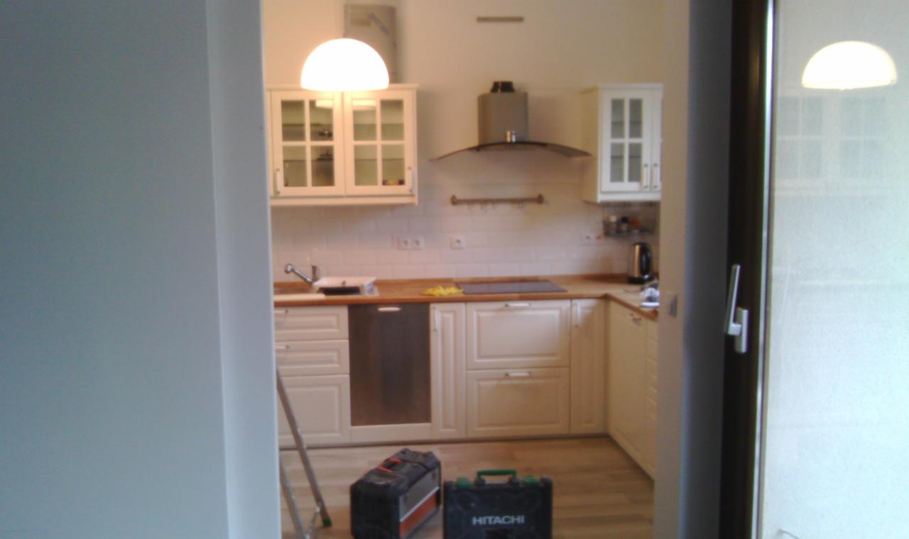 Jasna kuchnia po remoncie, widoczna przez otwarte drzwi. Na podłodze skrzynki narzędziowe marki Hitachi, oparta drabina. Białe szafki wiszące i stojące, drewniany blat, srebrny okap.