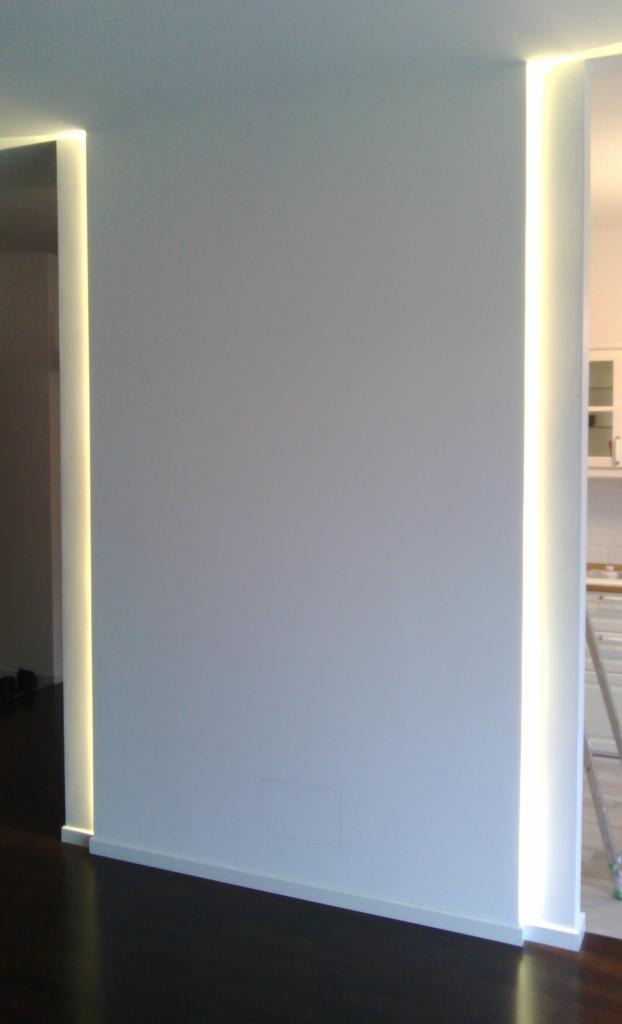 Biała ściana z wbudowanym oświetleniem LED w pionowych szczelinach, widoczny fragment ciemnej podłogi i fragment kuchni w tle.