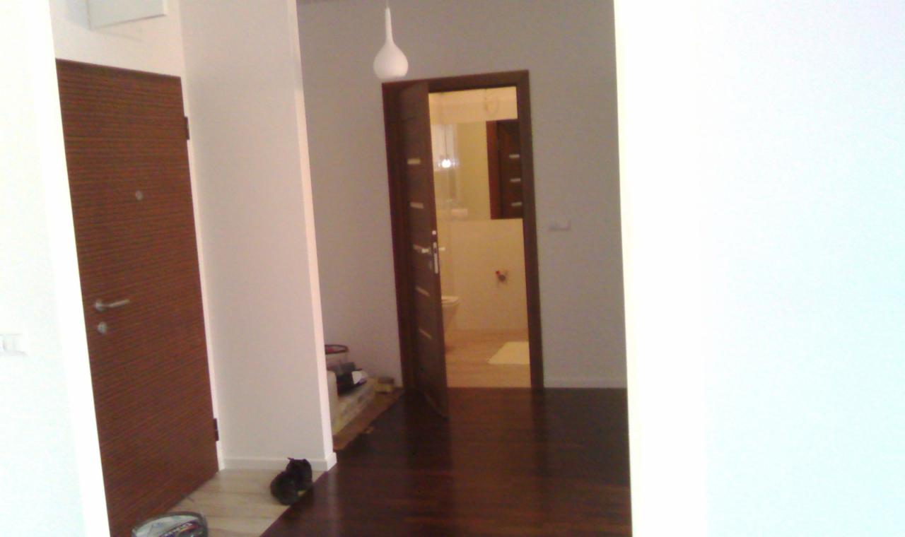 Widok na korytarz z dwiema parami drzwi, jedne wejściowe drewniane z wizjerem, drugie otwarte do łazienki z widoczną toaletą i białą umywalką.