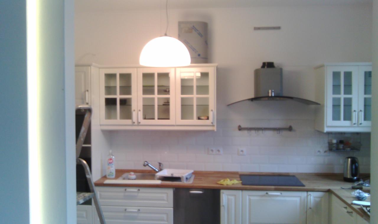 Wnętrze kuchni w trakcie remontu, widoczne białe szafki, drewniany blat, okap i lampa, na blacie leżą narzędzia i akcesoria.