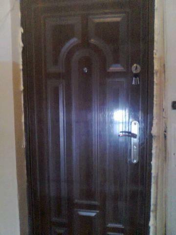 Ciemnobrązowe, metalowe drzwi wejściowe z ozdobnymi panelami i srebrną klamką, widoczne klucze w zamku, ściana po lewej stronie z widocznymi śladami po remoncie.